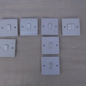 Op de afbeelding: Acht witte lichtschakelaars met een enkele of dubbele schakelaar. De schakelaars zijn gemonteerd op een lichtgrijze ondergrond.