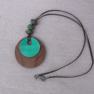 Puede incluir: Un collar de madera marrón con un círculo verde en el centro. El collar tiene un cordón de cuero marrón y cuentas plateadas.