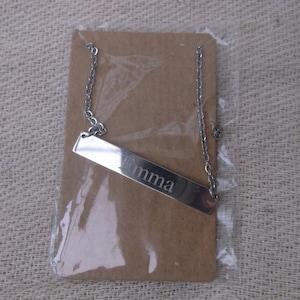 May include: A silver-toned metal bar necklace featuring the name Emma engraved in a serif font. The necklace is attached to a rectangular brown cardboard display card and is enclosed within a clear plastic protective sleeve for storage.