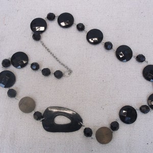 以下が含まれることがあります： 丸型と楕円形のビーズが付いた黒いネックレス。ネックレスは黒と茶色のビーズが混ざっており、銀色のチェーンでつながっています。