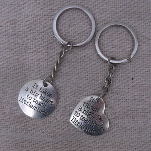 May include: Two silver keyrings, one round and one heart-shaped, both engraved with the phrase "It takes a big heart to teach little minds". The heart-shaped one has an apple graphic.