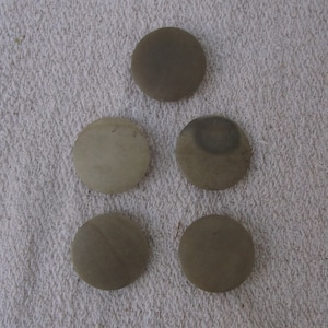 May include: Five round, smooth, light brown stones arranged in a triangular pattern on a white textured surface.