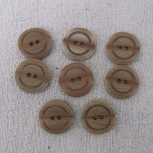 Puede incluir: Una colección de ocho botones redondos de color marrón claro con dos agujeros cada uno. Cada botón tiene un diseño de cara sonriente tallada. Los botones están dispuestos sobre una superficie texturizada blanquecina.