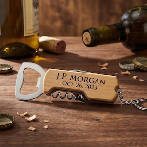 May include: A wooden bottle opener and corkscrew combination with the engraved text "J.P. MORGAN OCT. 26, 2023". The opener has a silver metal bottle opener and corkscrew. The item is on a wooden surface with wine-related items.