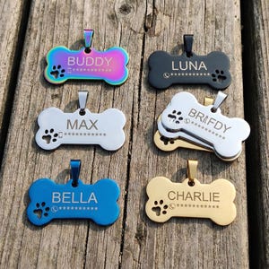 May include: A collection of bone-shaped pet ID tags in various colors, including iridescent, black, silver, blue, and gold. Each tag features a pet's name and contact information, along with paw print and phone icons. The tags are displayed on a wooden surface.