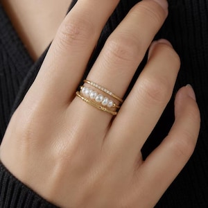 Könnte beinhalten: Ein Stapel aus drei goldenen Ringen an einem Finger. Der oberste Ring hat ein schmales Band mit kleinen eingebetteten Steinen. Der mittlere Ring zeigt fünf kleine weiße Perlen in einer Reihe. Der untere Ring hat ein strukturiertes Goldband.