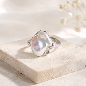 Peut inclure: Une bague en argent ornée d'une grande perle baroque irrégulière et iridescente en son centre. L'anneau métallique présente une finition organique texturée rappelant l'écorce d'un arbre, enserrant la perle dans un sertissage à griffes.