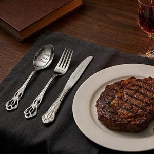 Peut inclure: Un ensemble de couverts argentés à trois pièces avec des noms gravés, comprenant une cuillère, une fourchette et un couteau, à côté d'une assiette avec un steak grillé. La cuillère porte l'inscription "Emily Laura Anna".