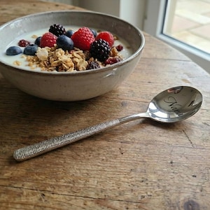 May include: A stainless steel spoon with the name "Nava" engraved on the bowl, next to a bowl of yogurt, granola, and mixed berries. The spoon has a textured handle. The bowl is light gray and sits on a wooden surface.