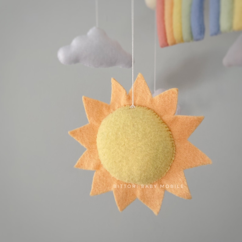 May include: A felt sun mobile with a yellow center and orange rays. The sun is hanging from a white string. The text "Bitttori Baby Mobile" is visible at the bottom of the image.