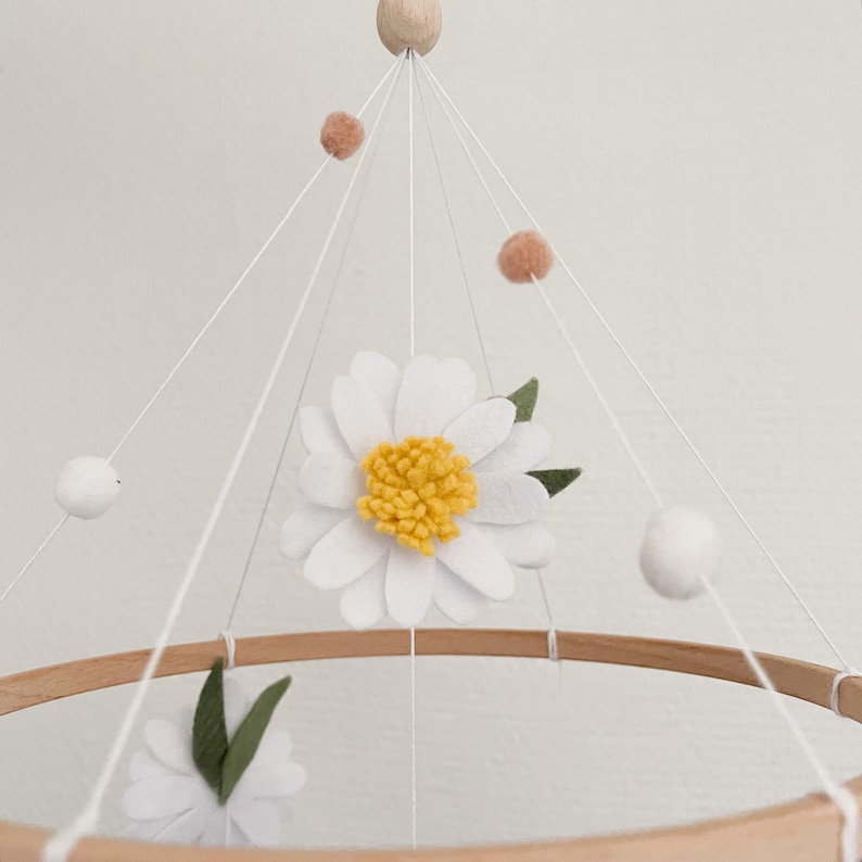 May include: A white and yellow felt daisy flower mobile with white pom poms and peach felt balls hanging from a wooden ring.