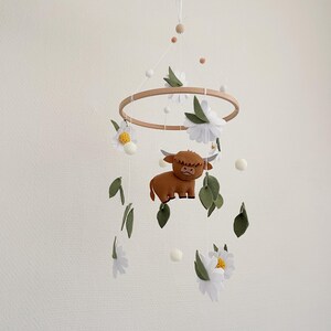 May include: A wooden baby mobile with a brown felt highland cow, white felt daisies, and green felt leaves. The mobile hangs from a white ceiling.
