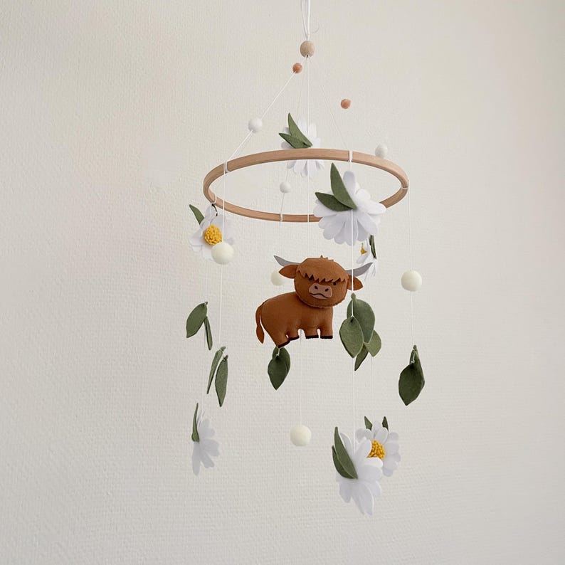 May include: A wooden baby mobile with a brown felt highland cow, white felt daisies, and green felt leaves. The mobile hangs from a white ceiling.