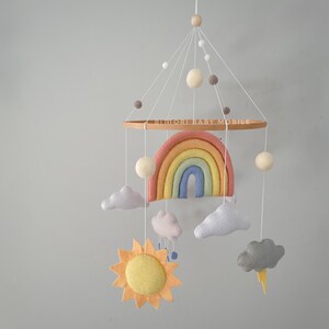 May include: A wooden baby mobile with a rainbow, clouds, sun, and raindrops. The mobile is made of felt and has a wooden ring with the text "Blumori Baby Mobile".