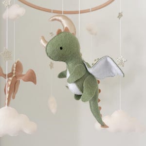 May include: A green felt dragon with white wings and a gold moon hanging from a wooden mobile. The dragon has a brown felt tail and a white felt belly. The mobile also includes white felt clouds and gold glitter stars.