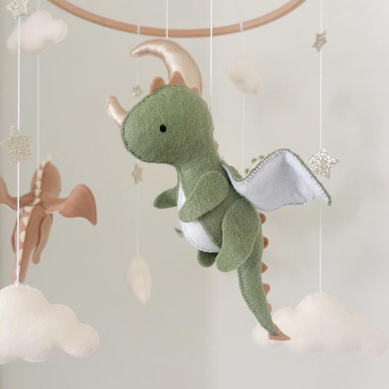 May include: A green felt dragon with white wings and a gold moon hanging from a wooden mobile. The dragon has a brown felt tail and a white felt belly. The mobile also includes white felt clouds and gold glitter stars.