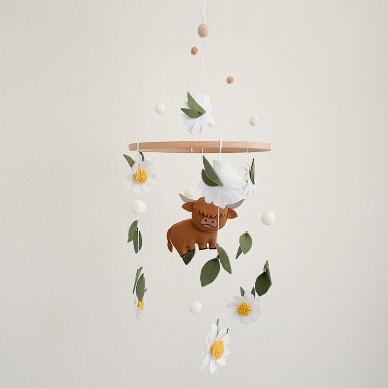 May include: A wooden baby mobile with a brown felt highland cow, white felt daisies, and green felt leaves. The mobile hangs from a wooden ring.