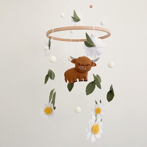 May include: A white and brown felt baby mobile with a highland cow, white flowers, and green leaves hanging from a wooden ring.
