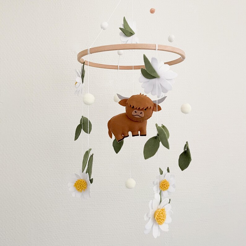 May include: A white and brown felt baby mobile with a highland cow, white flowers, and green leaves hanging from a wooden ring.