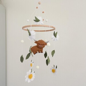 May include: A wooden baby mobile with a brown highland cow, white daisies, and green leaves. The mobile is hanging from a white ceiling.