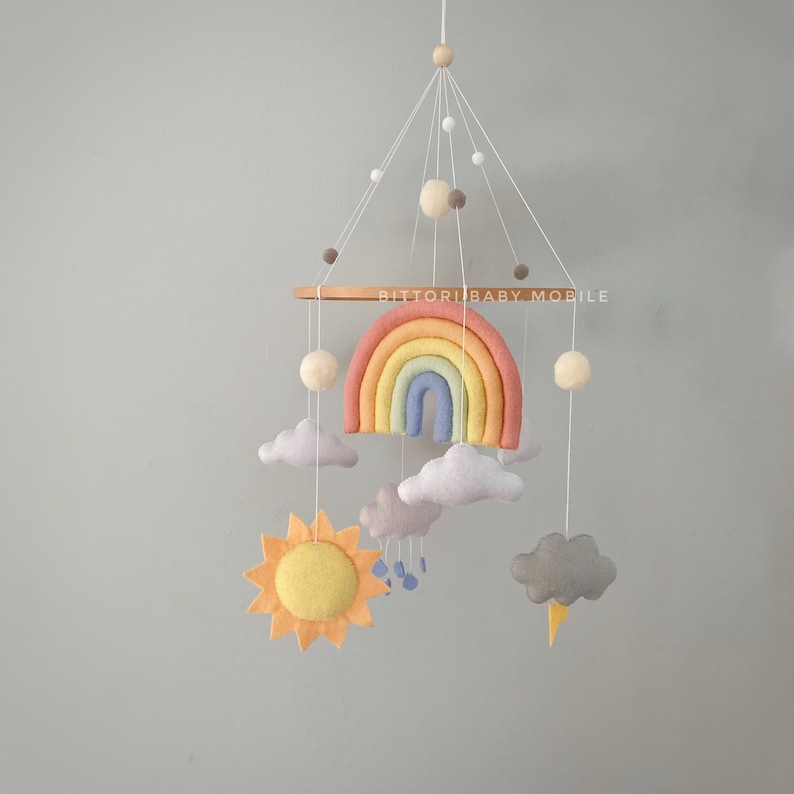 May include: A rainbow-themed baby mobile with a wooden ring, felt clouds, a sun, a rainbow, and raindrops. The mobile is hanging from a white ceiling.