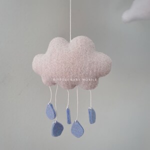 May include: A light pink felt cloud with blue felt raindrops hanging from it. The cloud has the text "Bitttori Baby Mobile" on it.