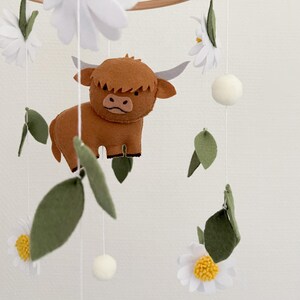 May include: A brown and white felt baby mobile with a highland cow, white flowers, and green leaves.