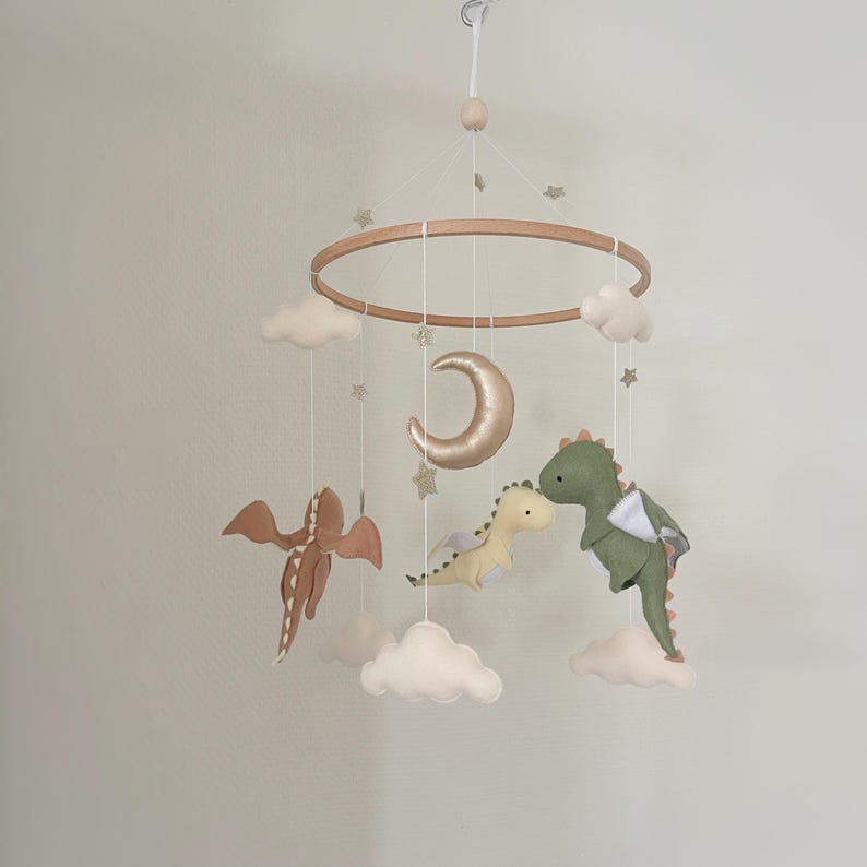 May include: A wooden baby mobile with a gold crescent moon, two felt dragons, and white clouds. The mobile is decorated with gold glitter stars.
