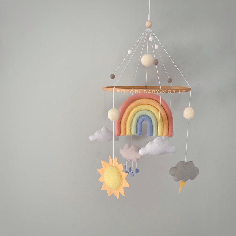 May include: A rainbow-themed baby mobile with a felt rainbow, clouds, sun, and raindrops. The mobile is suspended from a wooden ring with the text "Bitttori Baby Mobile".
