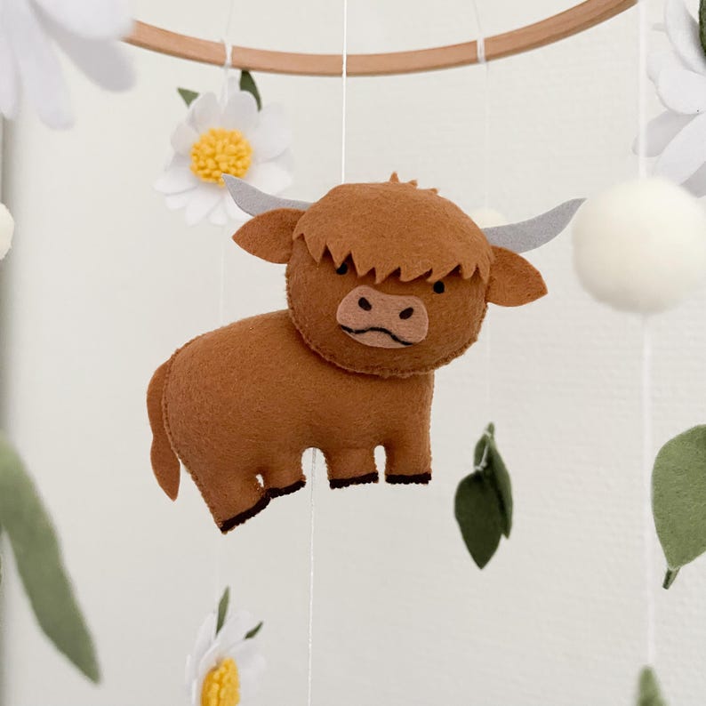 May include: A brown felt highland cow with a white felt nose and black felt eyes hangs from a wooden ring. The cow is part of a mobile with white felt flowers and green felt leaves.