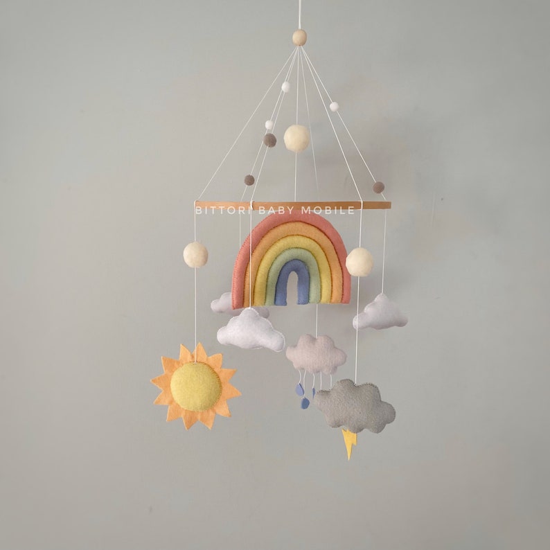 May include: A rainbow baby mobile with a rainbow, clouds, sun, and raindrops. The mobile is made of felt and has a wooden dowel with the text "BITTOR BABY MOBILE".