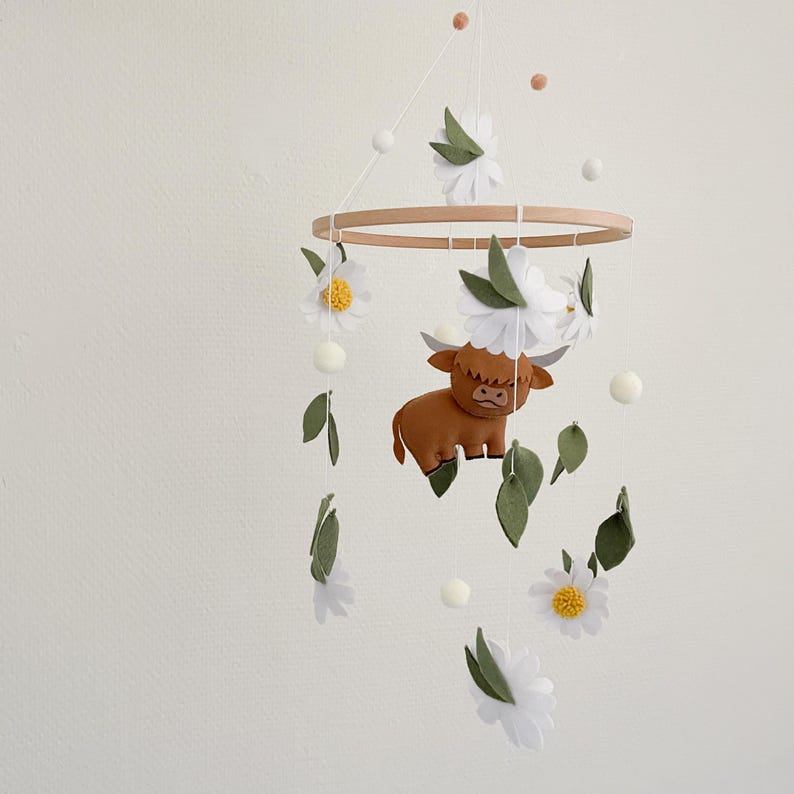 May include: A white and green felt baby mobile with a brown highland cow, white flowers, and green leaves hanging from a wooden ring.