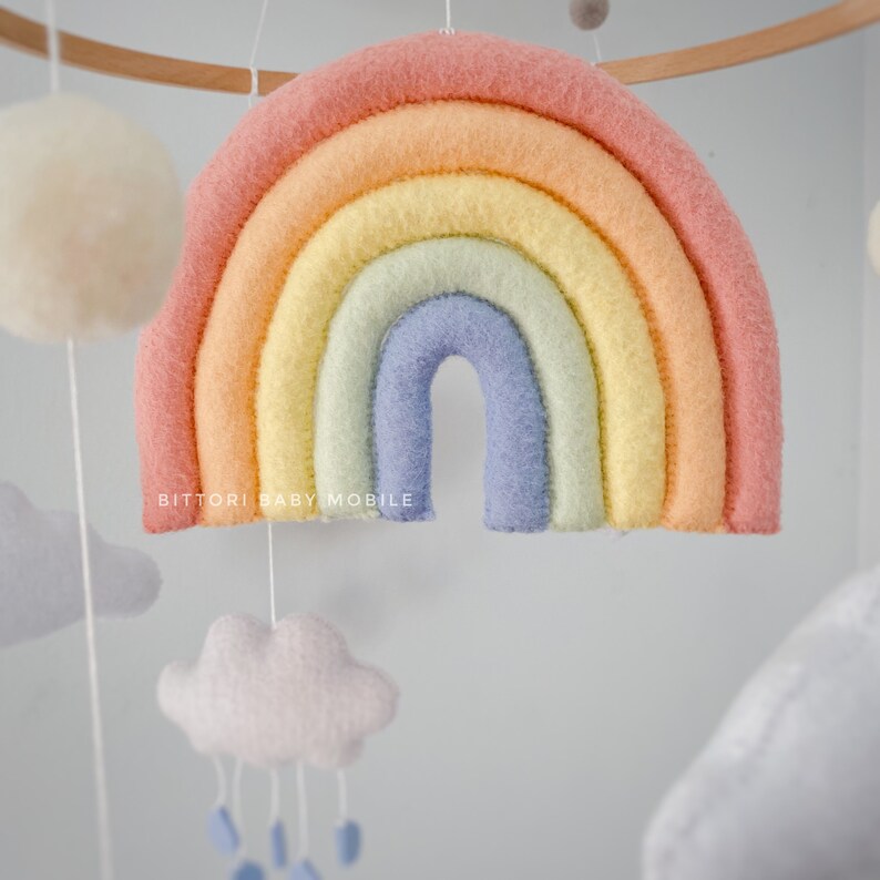 May include: A pastel rainbow mobile with a light blue, yellow, green, orange, and pink color scheme. The rainbow is made of felt and has a fluffy texture. The mobile also includes white felt clouds and blue felt raindrops.