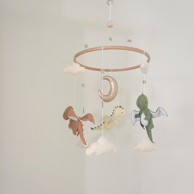 May include: A wooden baby mobile with a crescent moon, clouds, and three felt dragon toys. The dragons are green, brown, and cream.