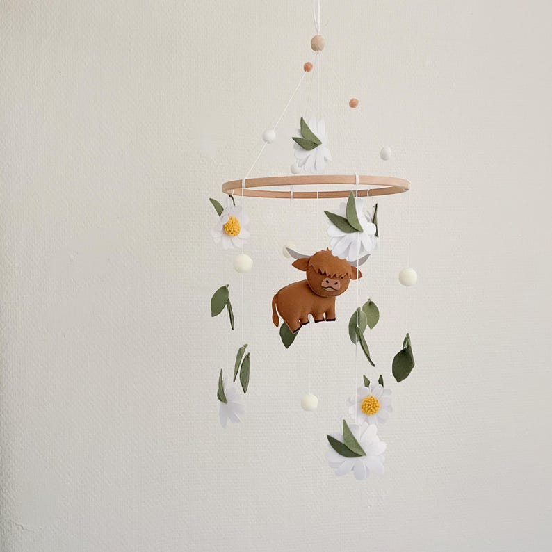 Handmade felt baby mobile featuring a cute Highland cow, white daisies, green leaves, and felt balls accents, hanging from a wooden hoop. Perfect rustic farm-themed nursery decor for a baby’s crib.