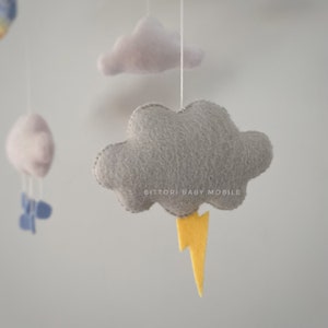 May include: A gray felt cloud with a yellow lightning bolt hanging from a white string. The cloud is part of a baby mobile with the text "Bitttori Baby Mobile" printed on it.