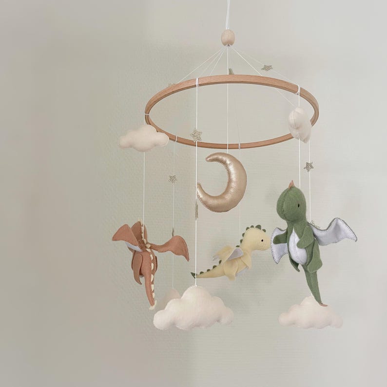 May include: A wooden baby mobile with a gold crescent moon, three felt dragons in green, yellow, and brown, and white clouds. The mobile is hanging from a white ceiling.
