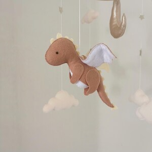 May include: A brown felt dragon with white wings hangs from a mobile. The dragon has a small black eye and a yellow felt outline. The mobile also includes white felt clouds.