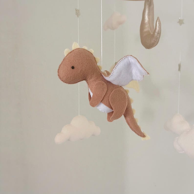 May include: A brown felt dragon with white wings hangs from a mobile. The dragon has a small black eye and a yellow felt outline. The mobile also includes white felt clouds.