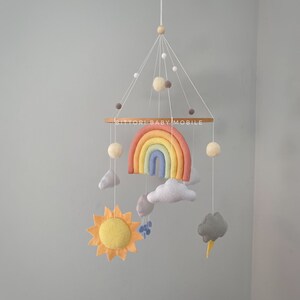 May include: A felt rainbow baby mobile with a yellow sun, gray clouds, and white pom-poms. The mobile is hanging from a wooden ring with the text "Bittorl Baby Mobile".
