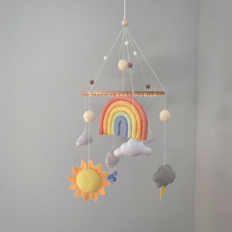 May include: A felt rainbow baby mobile with a yellow sun, gray clouds, and white pom-poms. The mobile is hanging from a wooden ring with the text "Bittorl Baby Mobile".