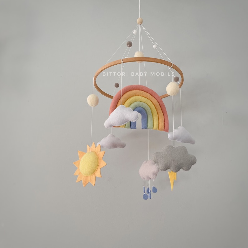 May include: A rainbow baby mobile with a wooden ring, felt clouds, a sun, a rainbow, and raindrops. The mobile is hanging from the ceiling and says "Bitttori Baby Mobile".