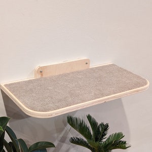 May include: A wall-mounted cat shelf with a gray fabric surface. The shelf is made of wood and has a rounded edge. There are two plants in pots on the floor below the shelf.