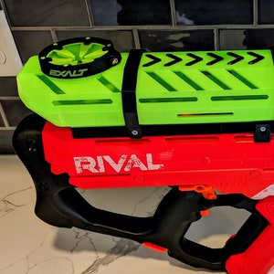 May include: A red and black Nerf Rival blaster with a green and black magazine attachment. The magazine has a black strap and the text "EXALT" on the top.