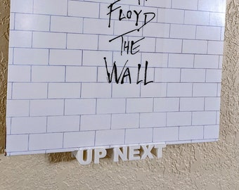 May include: A white vinyl record with the words "Pink Floyd The Wall" written in black marker on the front. The record is displayed on a white wall with a white plastic record holder that says "Up Next" in black letters.