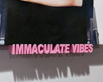 May include: A pink 3D plastic sign with the text "IMMACULATE VIBES" on a white wall. The sign is attached to a black and white poster with a parental advisory label.