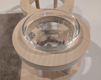 May include: A wooden pet feeder with two glass bowls. The feeder has a raised platform with a textured surface and a cutout for a water bowl.