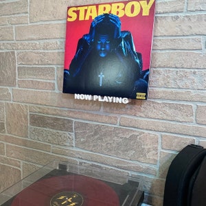 May include: A record player with a red vinyl record on the turntable. The record is playing "Starboy" by The Weeknd. A framed album cover for "Starboy" is hanging on the wall above the record player. The album cover features a man in a black leather jacket and a cross necklace. The text "NOW PLAYING" is printed on a white sign below the album cover.