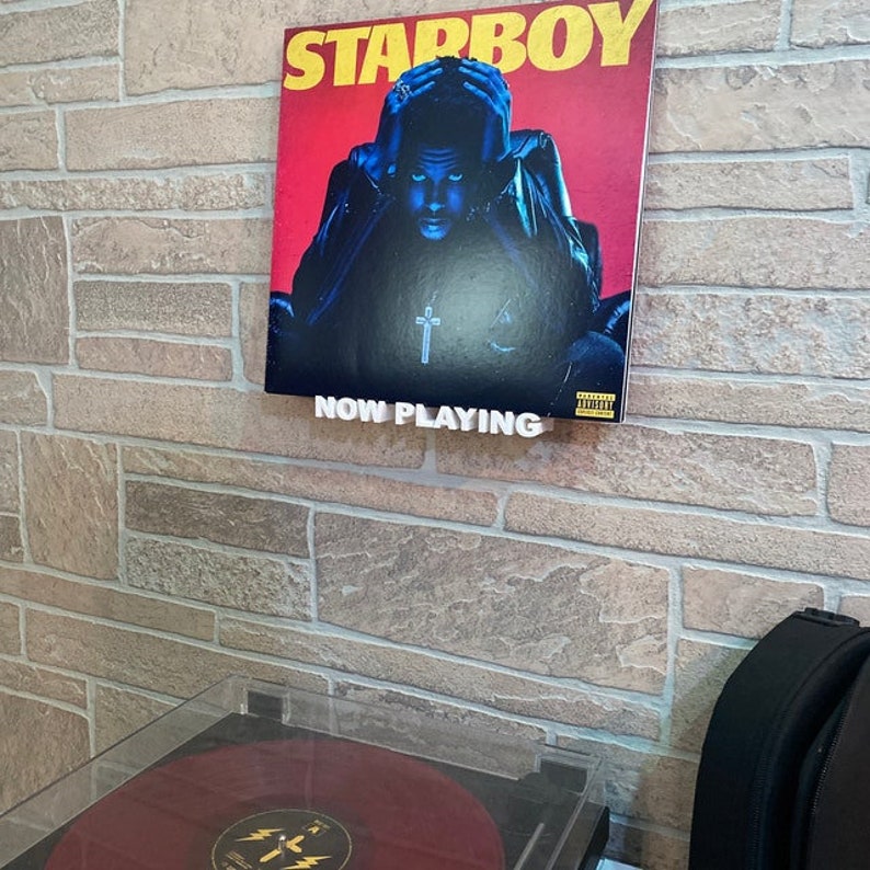 May include: A record player with a red vinyl record on the turntable. The record is playing "Starboy" by The Weeknd. A framed album cover for "Starboy" is hanging on the wall above the record player. The album cover features a man in a black leather jacket and a cross necklace. The text "NOW PLAYING" is printed on a white sign below the album cover.