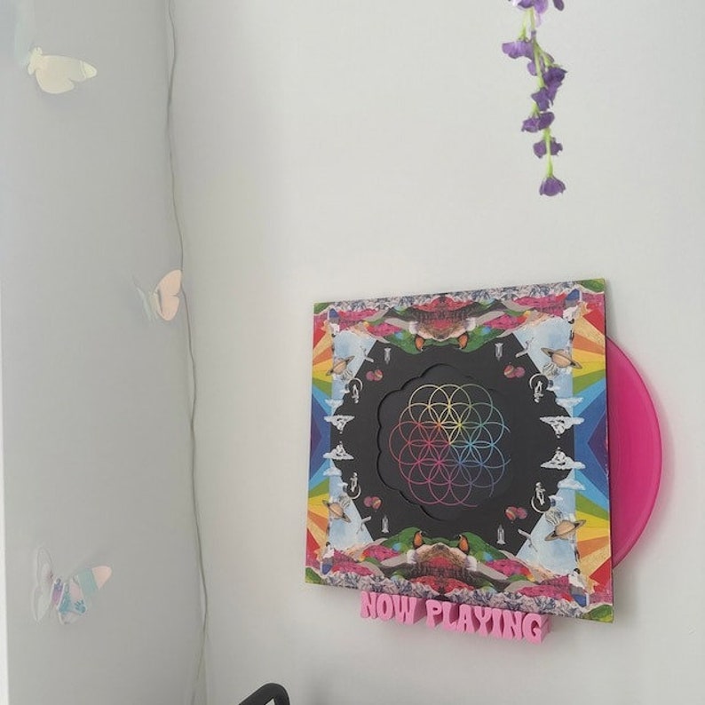 May include: A pink record player stand with a black and colorful record on it. The record has a rainbow mandala design and the text "NOW PLAYING" is written in pink letters below the record.